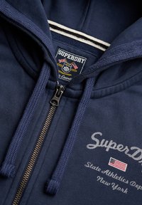 Marineblå Superdry hættetrøje med lynlås, snore, mærket lynlås, broderet logo og amerikansk flagpatch på brystet.