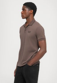 Mann som står i profil iført en mauve polo med EA7-logo og svarte jeans, med den ene hånden i lommen mot en enkel lys bakgrunn.