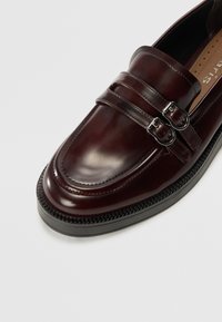 Burgundy leather loafers with a glossy finish, featuring dual silver buckles and black rubber sole with textured edge.