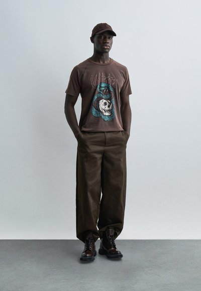 Άνδρας που φορά καφέ καπέλο, γραφικό μπλουζάκι με φίδι και κρανίο, φαρδιά καφέ παντελόνια και σκούρα δερμάτινα μποτάκια, στέκεται με τα χέρια στις τσέπες.