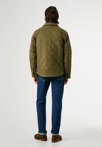 Gequiltede olijfgroene jas met een kraag, drukknopen en gestructureerde stof, gecombineerd met donkerblauwe spijkerbroek en bruine schoenen.