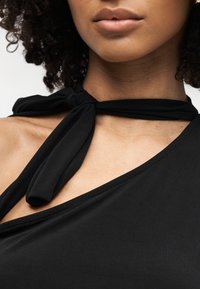 Γυναίκα που φοράει μαύρο τοπ με έναν ώμο και δεμένο λουρί γύρω από το λαιμό της, αποκαλύπτοντας το κλειδικό οστό και μέρος των χειλιών και του πηγουνιού της.