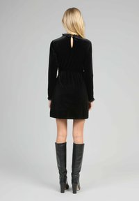 Femme portant une robe mini en velours noire à manches longues avec une petite ouverture en forme de clé au dos, associée à des bottes noires à talons jusqu'au genou.