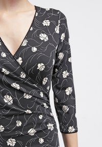 Zwarte wikkeljurk met driekwartmouwen, voorzien van een bloemenpatroon in crème, gestructureerde stof en gegolfde afwerking langs de halslijn.