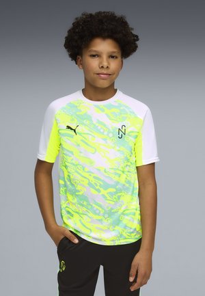 Ragazzo con capelli ricci in piedi con una mano in tasca, indossa una maglietta sportiva a motivo neon giallo e bianco e pantaloncini neri su sfondo grigio.