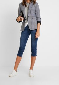 Grå och blårutig blazer med svarta lädermanschetter. Matchad med denimbukser i capri-längd och vita sneakers, håller en svart clutch.