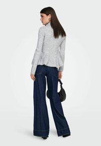 Šedý peplum svetr s dlouhými rukávy, texturovanou tkaninou a vysokým výstřihem, kombinovaný s tmavě modrými džínami se širokými nohavicemi a černou kabelkou.