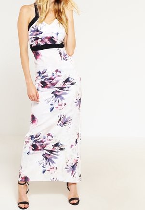 Femme portant une robe maxi blanche sans manches avec des imprimés floraux violets et roses et des talons noirs à bride à la cheville, debout sur un fond uni.
