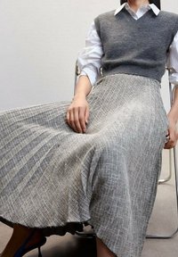 Gilet grigio lavorato a maglia sopra una camicia bianca, abbinato a una gonna grigia plissettata con un motivo testurizzato e un orlo ondulato. Scarpe con tacco alto blu visibili.