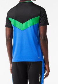 Man med kortärmad sportskjorta i svart, grön och blå färgblock och svarta shorts med en sidologotyp.