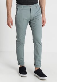 Mężczyzna w jasnoszarych slim-fit spodniach z podwiniętymi nogawkami i czarnych trampkach, stojący na tle białego tła.