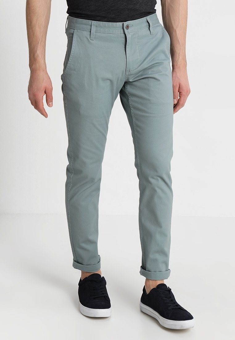 Mężczyzna w jasnoszarych slim-fit spodniach z podwiniętymi nogawkami i czarnych trampkach, stojący na tle białego tła.