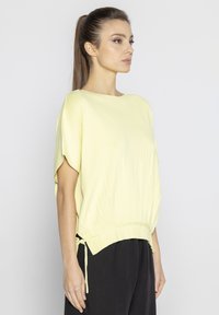 Jasnożółta bluzka z krótkim rękawem, z okrągłym dekoltem i ribbowym wykończeniem, z regulowanymi bocznymi troczkami i luźnym krojem.