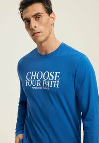 Man wearing a blue long-sleeve shirt with "CHOOSE YOUR PATH HENDERSON STUDIOS" text, posing against a plain light background.