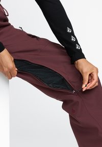 Burgundy vattentåliga byxor med dragkedjeficka på sidan, meshnätfoder och justerbar dragsko. Svart långärmad topp med logotypdetaljer.