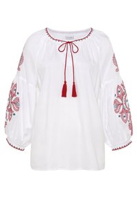 Blouse blanche avec broderie florale rouge sur les manches, encolure élastique avec cordon et pompons, et accents décoratifs bleus.