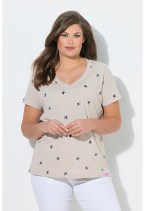 Kvinde med langt brunt hår iført beige T-shirt med V-hals med små sorte søstjerner og hvide bukser, stående mod en hvid baggrund.