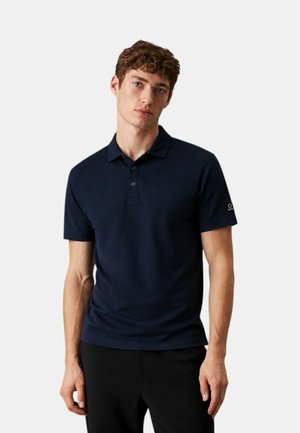 Giovane uomo con capelli castani ricci che indossa una polo blu navy a maniche corte e pantaloni neri, in piedi su uno sfondo semplice.