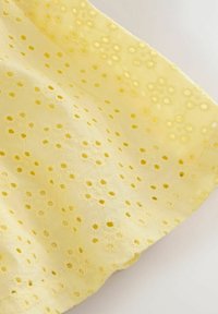 Tissu jaune avec de petites fleurs brodées et des motifs de découpes circulaires, présenté avec des plis doux sur un fond clair.