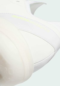 Bijela atletska cipela s glatkom kožnom gornjom dijelu, prozirnom gumenom potplatu i "ADIZERO INSTINCT+" oznakama u neonski žutim naglascima.