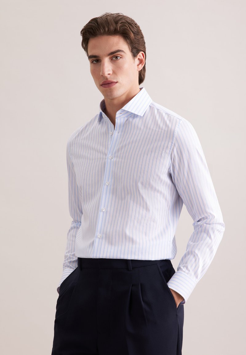 Homme portant une chemise à rayures bleu clair et blanc, boutonnée et rentrée dans un pantalon plissé noir, les mains dans les poches, fond neutre.