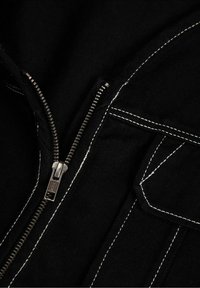 Närbild av ett delvis oöppnat svart plagg med silverfärgad dragkedja och vit kontrastsömnad som omger en ficklock.