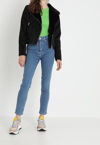 Chaqueta de moto de ante sintético negra, suéter de punto verde, jeans skinny azules y zapatillas beige con detalles en gris y calcetines amarillos.