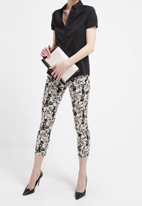 Camicia nera a maniche corte con bottoni, pantaloni capri a stampa floreale, tacchi alti neri, che tiene in mano una borsa a tracolla piegata con un design testurizzato.