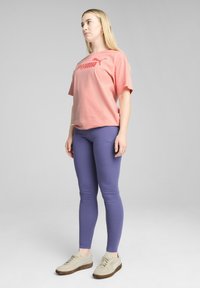 Puma majica s logotipom v koralni barvi, sproščen kroj; v kombinaciji z oprijetimi vijoličnimi leggins; svetle superge z teksturiranimi detajli.
