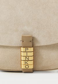 Borsa in suède beige con tracolla in pelle, caratterizzata da dettagli in metallo dorato che formano la scritta "PINKO" in verticale sulla chiusura a patta.