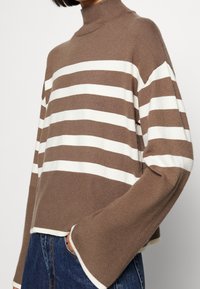 Kvinna iklädd en brun och vit horisontellt randig polo-tröja med långa, breda ärmar och blå jeans.