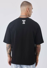 Camiseta negra de manga corta con un corte relajado, que presenta el texto "CAMBIA EL JUEGO" en letras blancas en negrita en la parte superior de la espalda.