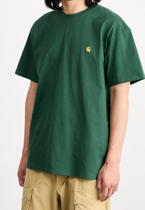 Osoba ubrana w ciemnozieloną koszulkę z krótkim rękawem z małym żółtym logo na piersi oraz beżowe szorty typu cargo, stojąca na białym tle.