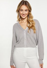 Cardigan grigio a maglia ribbed con scollatura a V, maniche lunghe e chiusura frontale con bottoni decorativi. Indossato sopra pantaloni bianchi.