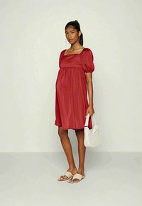 Mulher grávida vestindo um vestido vermelho com mangas bufantes, segurando uma bolsa branca, de pé descalça contra um fundo claro.