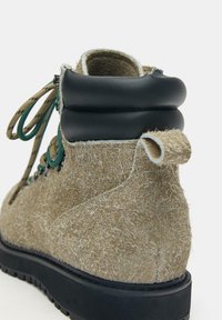 Zbliżenie beżowego, teksturowanego buta trekkingowego z czarną wyściełaną cholewką, zielono-beżowymi sznurowadłami oraz czarną gumową podeszwą.