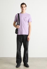 Camiseta de algodón en lavanda claro con logo negro, combinada con pantalones de mezclilla gris oscuro y zapatillas negras. Pequeño bolso negro cruzado sobre el cuerpo.