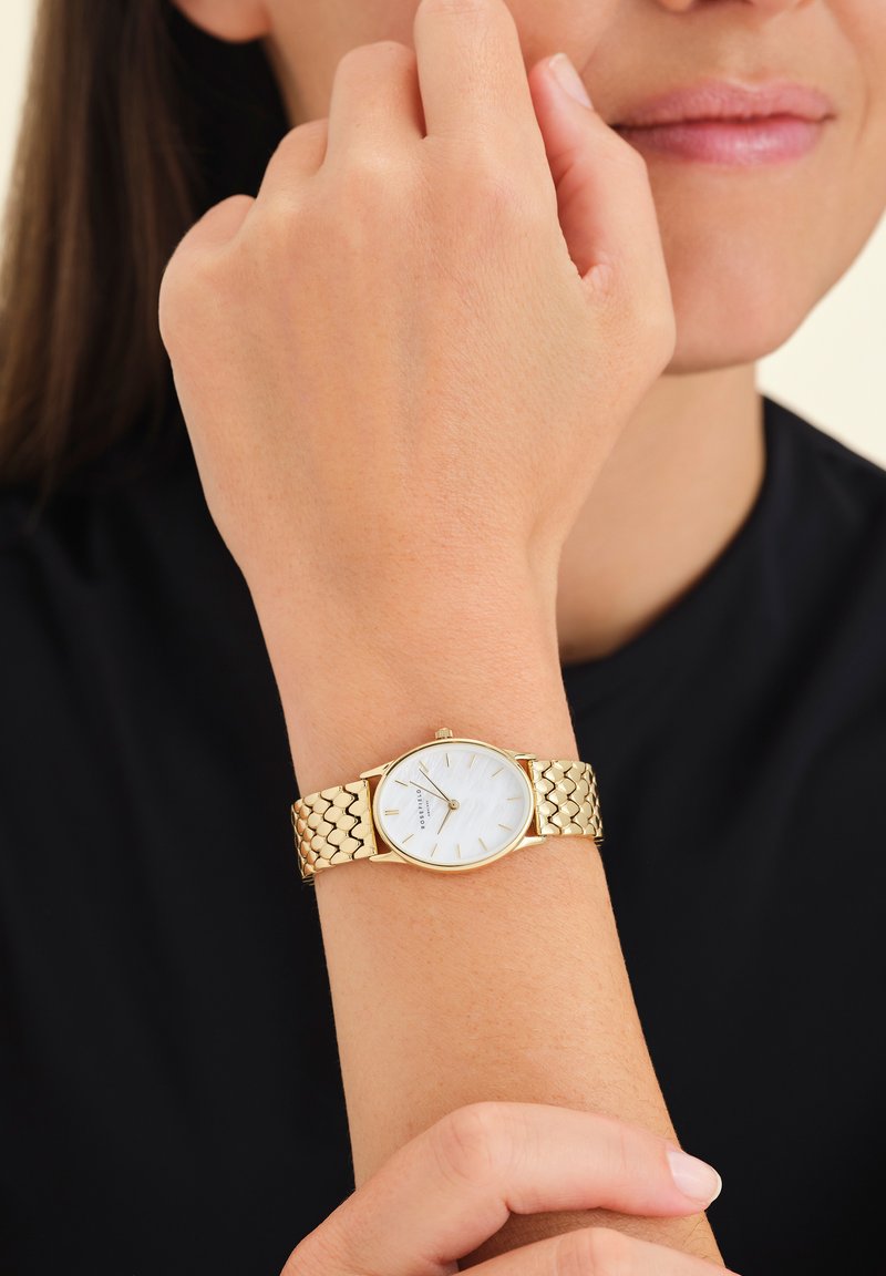 Orologio da polso dorato con bracciale texture, quadrante bianco rotondo e semplici indicatori delle ore. Indossato su un polso con un tono di pelle naturale.