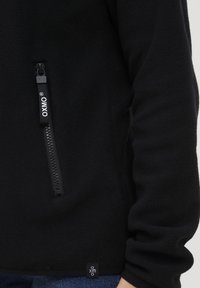 Svart fleece-pull med glidelåslomme, med teksturert stoff, rund nederkant, og et lite merketikett nederst.