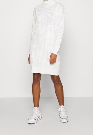 Woman wearing a white cable-knit turtleneck sweater dress paired with white high-top sneakers, standing against a plain background.
