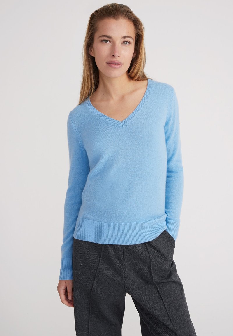 Pull léger bleu, en V, fabriqué en tissu doux. Présente des poignets et un ourlet côtelés, avec une coupe décontractée. Associé à un pantalon sombre et ample.