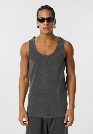 Grijze mouwloze tanktop met een geribbelde textuur, voorzien van een ronde halslijn en een rauw afgewerkte onderzijde. Gekozen in combinatie met zwarte zonnebril en een zilveren ketting.