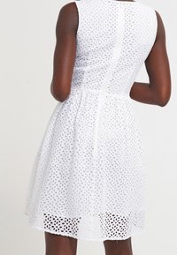 Biała sukienka bez rękawów wykonana z ażurowej tkaniny, z dopasowanym gorsetem i rozkloszowanym dołem. Charakterystyczne okrągłe wycięcia i podszewka na dole.