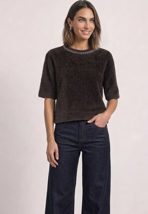 Femme aux cheveux mi-longs portant un haut court à manches courtes foncé et duveteux avec un col orné de perles et un jean large foncé, souriante, la main dans la poche.