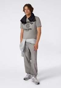 Mężczyzna w szarej koszulce "Champion Training Academy", jasno szarych spodniach, trampkach, z bluzą zawiązaną w talii i ręcznikiem na szyi, stoi.