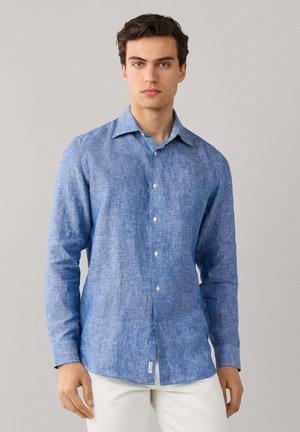 Junger Mann, der ein langärmeliges blaues Leinenhemd und weiße Hosen trägt, vor einem schlichten hellgrauen Hintergrund stehend.