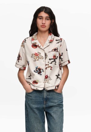 Kobieta z długimi ciemnymi włosami, ubrana w koszulę z krótkim rękawem zapinaną na guziki z kolorowymi abstrakcyjnymi nadrukami stworzeń i niebieskie dżinsy, ręce w kieszeniach.