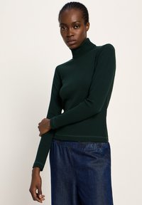 Frau mit geflochtenen Haaren, die einen dunkelgrünen, gerippten Rollkragenpullover und lockere dunkelblaue Jeans trägt, steht vor einem hellen Hintergrund.