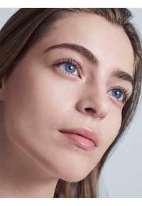 Close-up of a face with smooth, fair skin, blue eyes, defined brows, and lightly tinted lips; hair is straight and partially visible.
