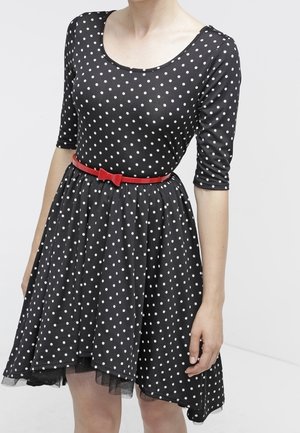 Femme portant une robe noire à pois blancs, manches trois-quarts, et une fine ceinture rouge avec un nœud à la taille.